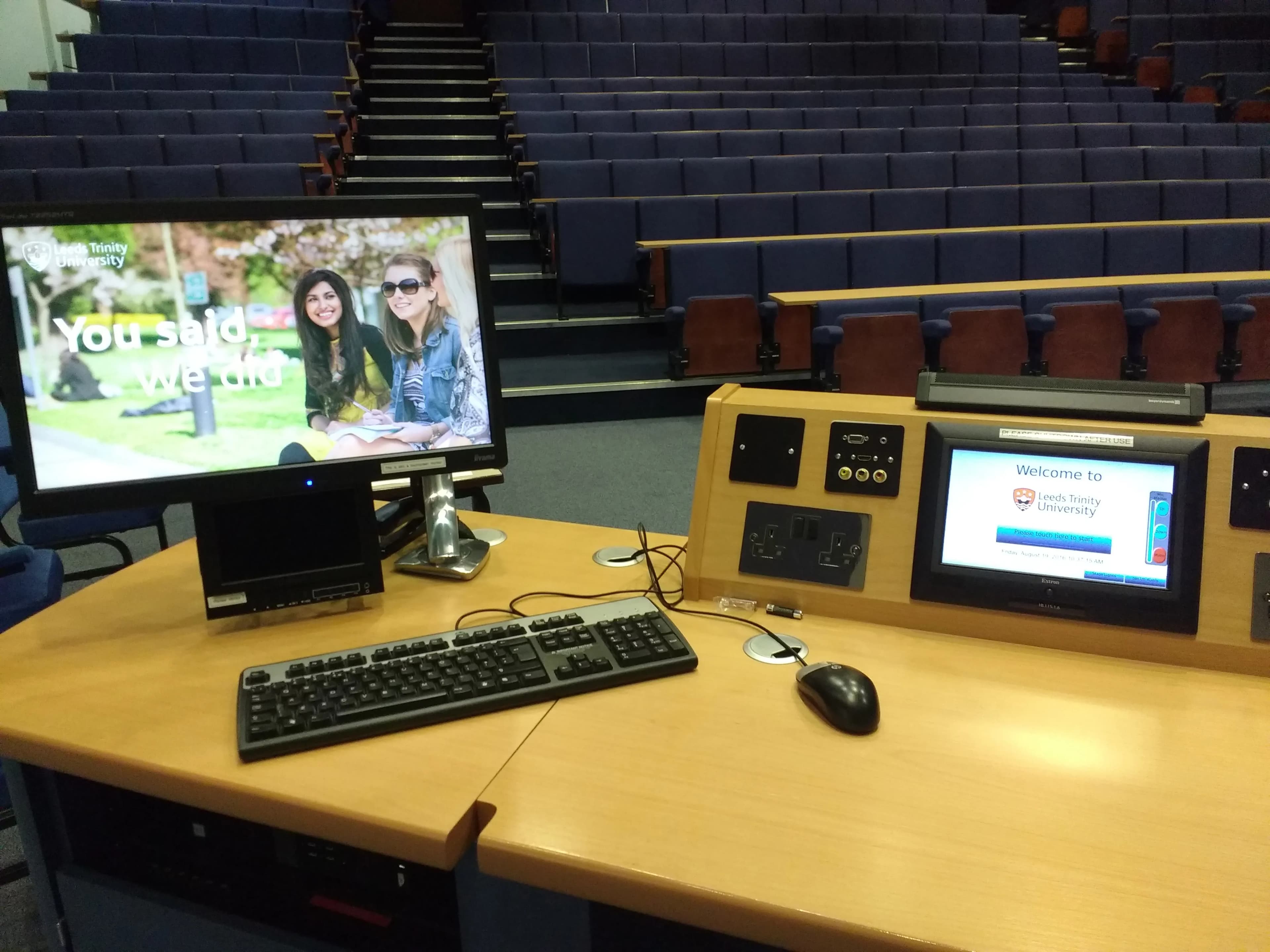 Computer equipment on a wooden desk facing empty tiered blue seats in a lecture hall.