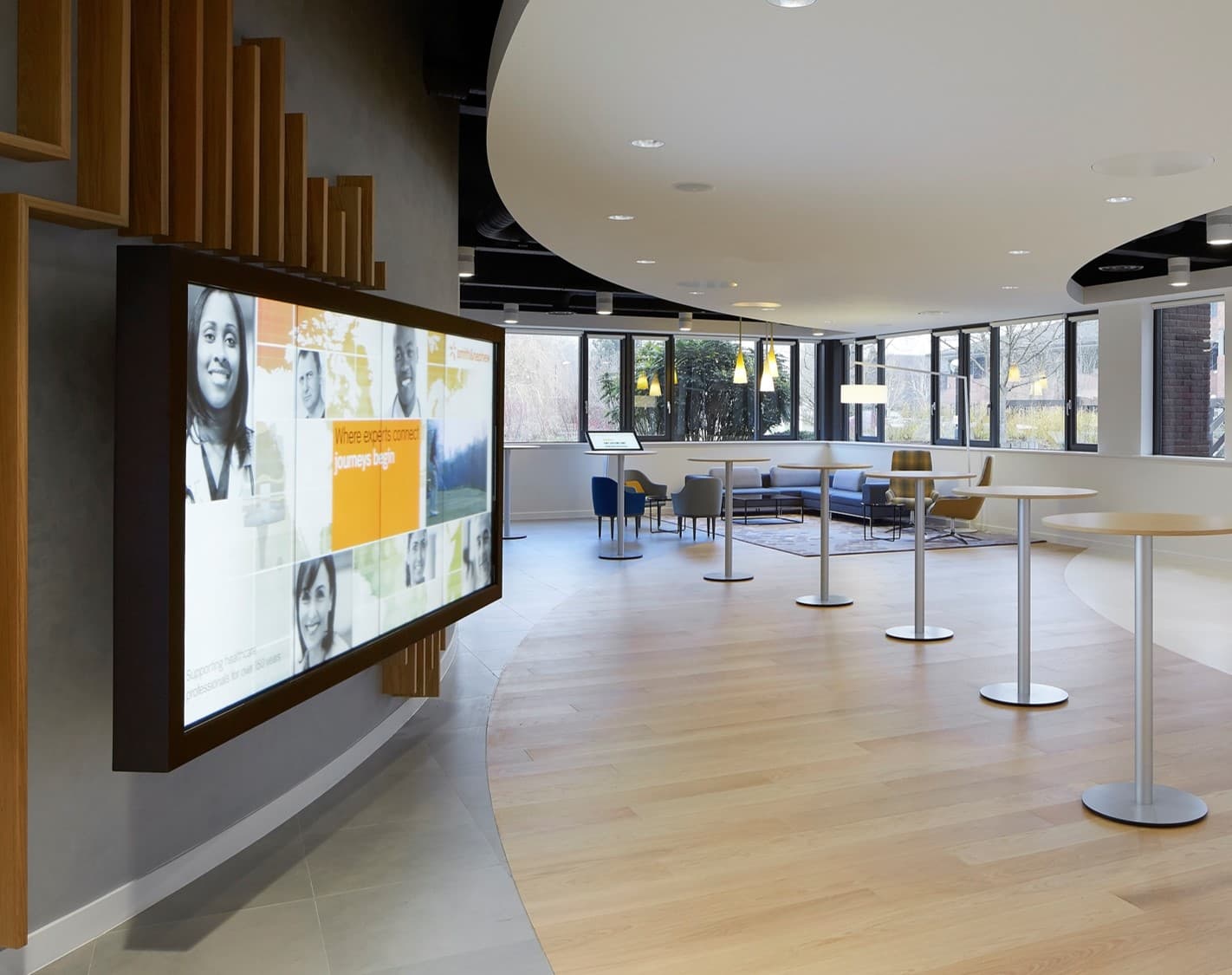 Modern office lobby featuring a large digital display, tall bistro tables, and lounge seating.